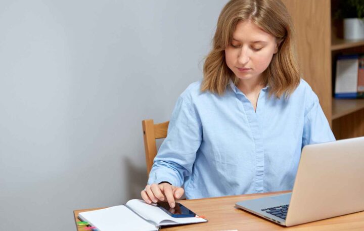 Young woman sitting at her desk looking at her phone and computer