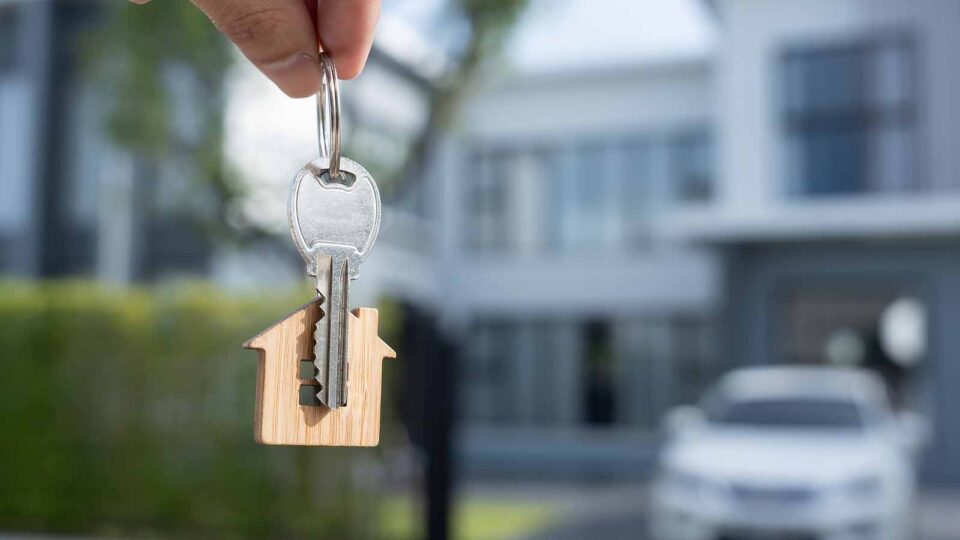 Keys to an investment property with a house in the background and a car parked