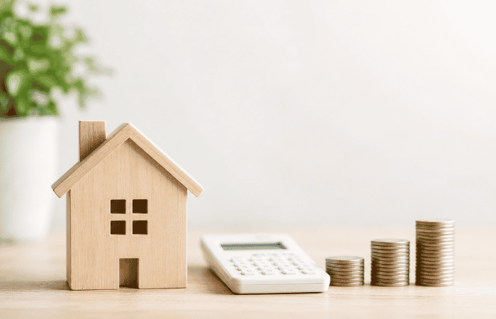 A neutral toy wooden house beside a calculator and stacked coins.